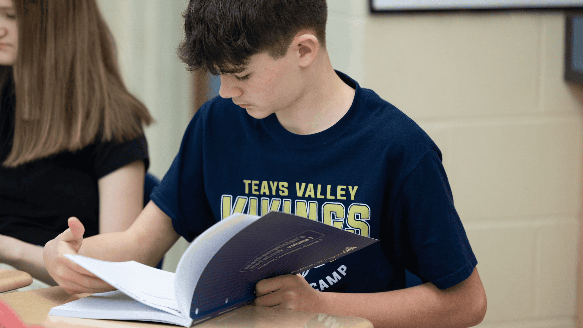 A student reading through their math workbook at a desk beside a classmate